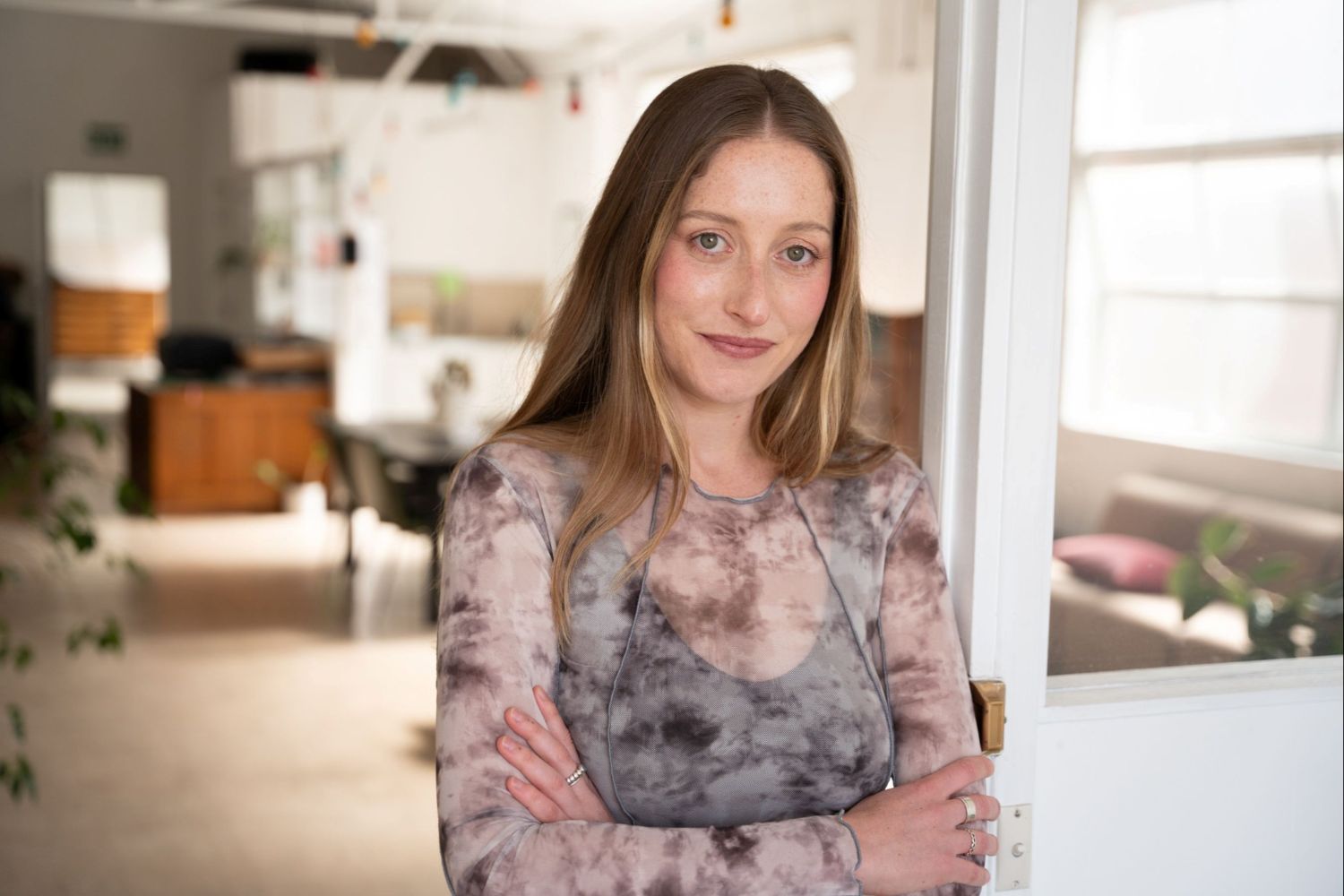 A woman wearing a grey shirt stands with her arms crossed in an office environment, smiling
